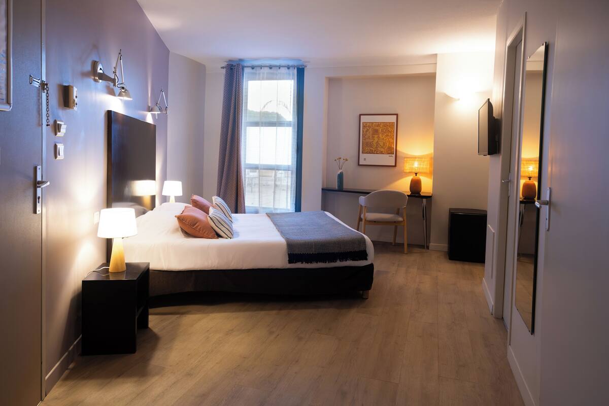 A hotel room with a bed, bedside lamps, a desk with a chair, a window with curtains, and a framed picture on the wall.