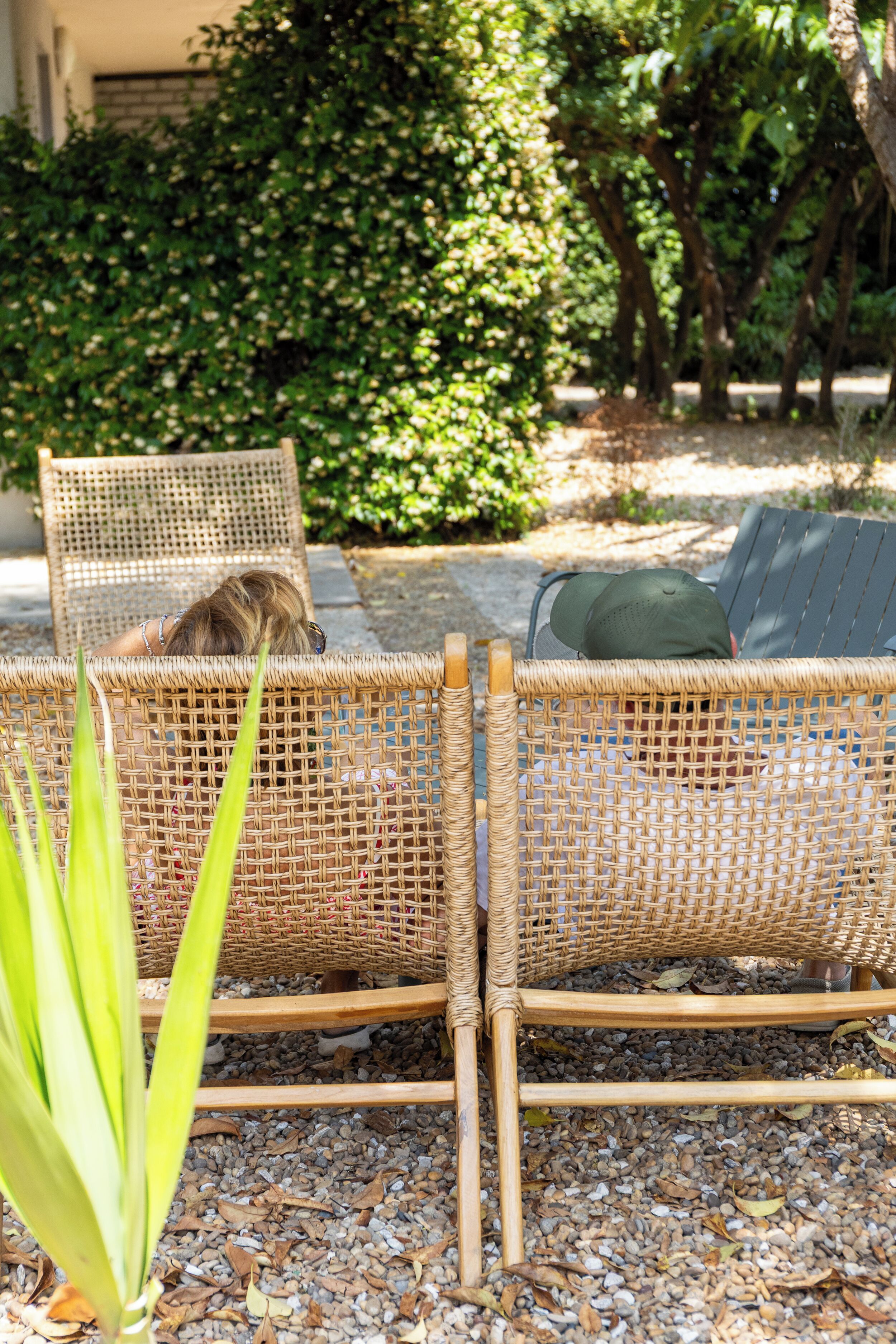 Wicker loungestoelen met kussens, geplaatst op een grindpad met een tuingedeelte op de achtergrond.