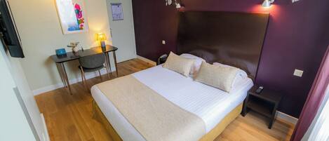A hotel room with a bed, bedside table, desk, chair, and a wall-mounted lamp.