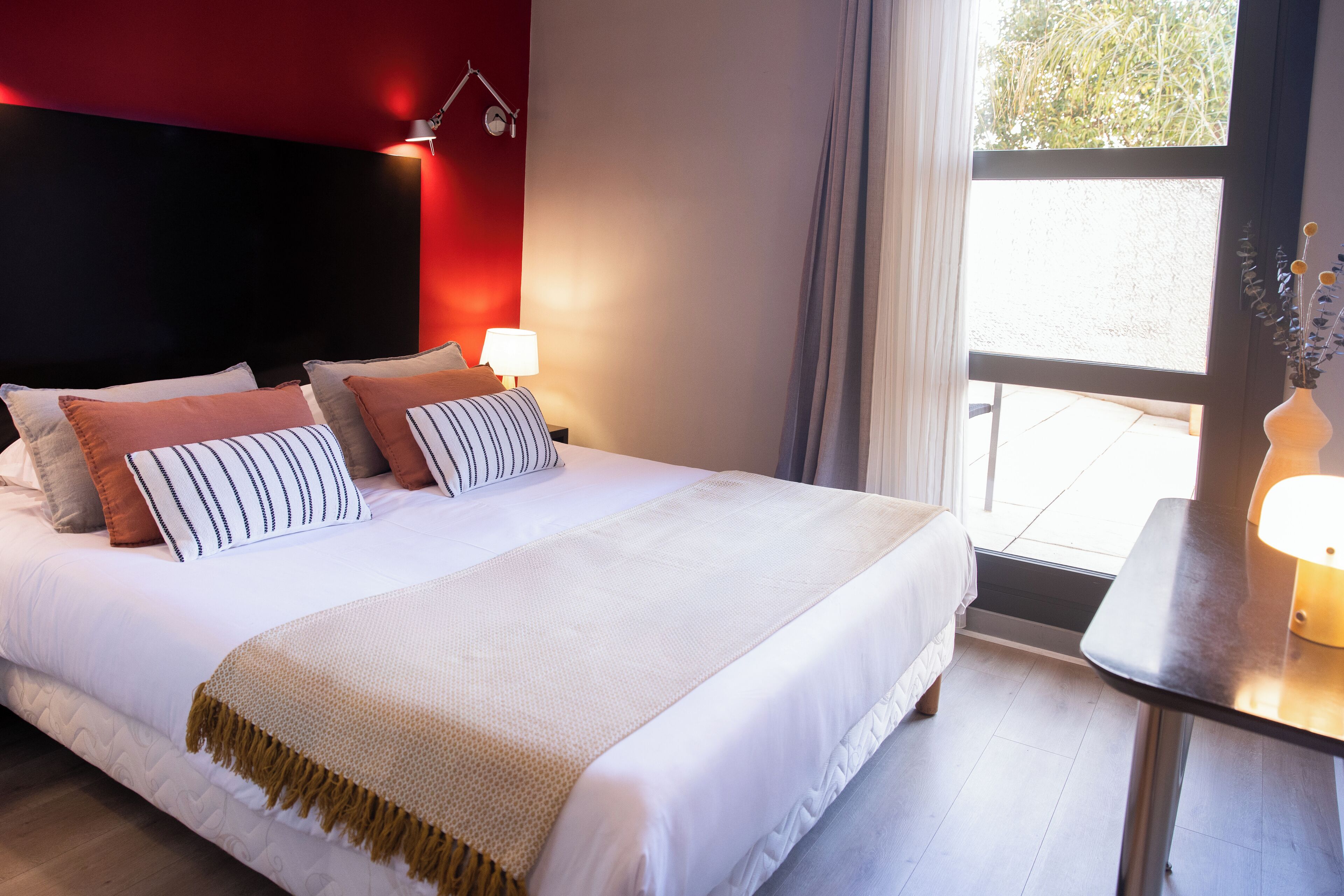 A neatly made bed with multiple pillows and a white blanket, a bedside table with a lamp, and a window with curtains.