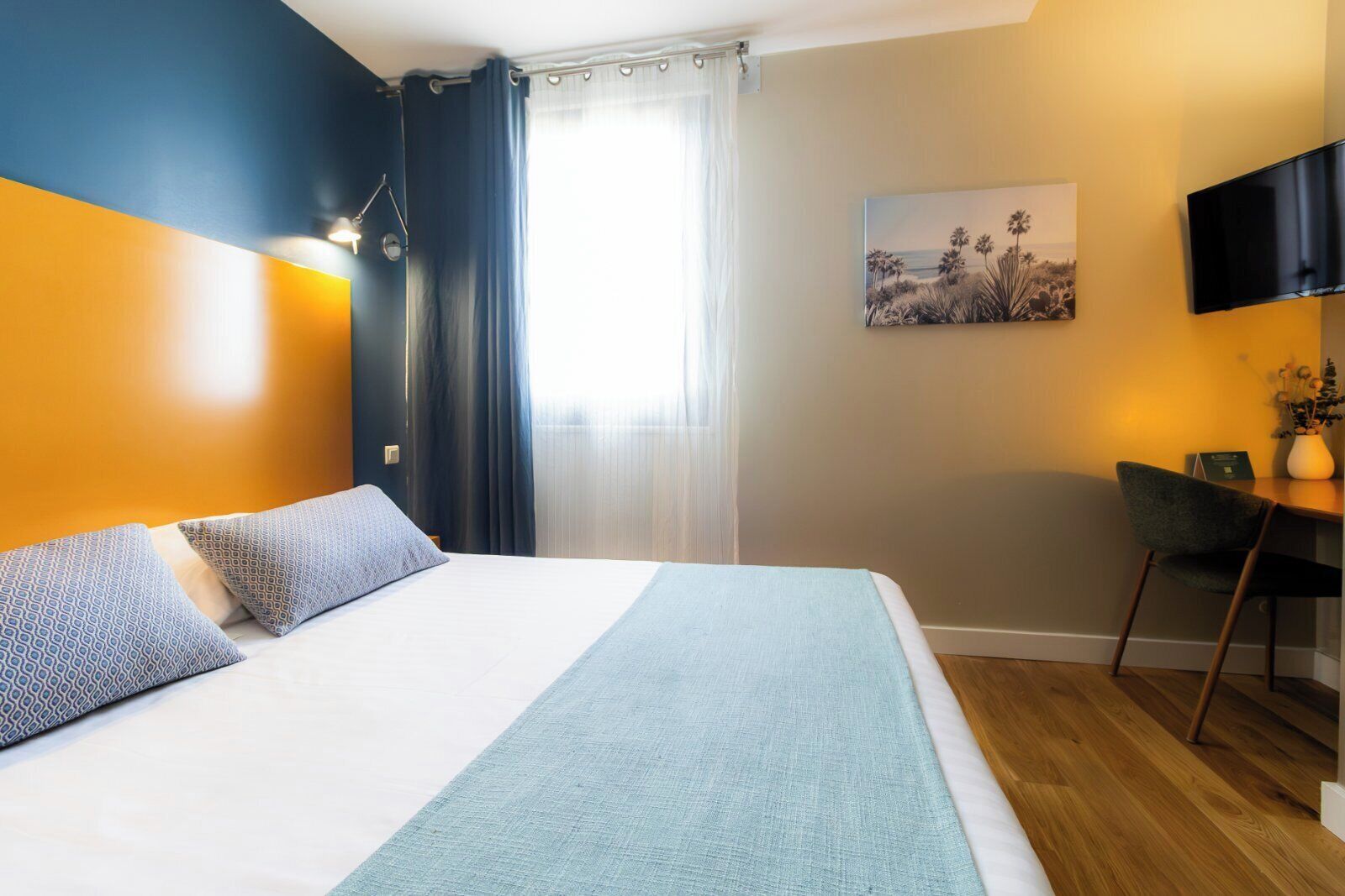 A hotel room with a bed, a desk, a chair, a TV, and a painting on the wall.