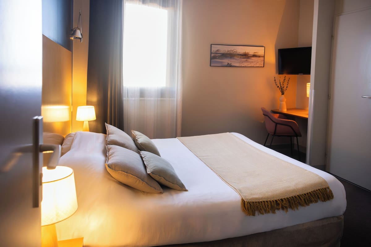 A hotel room with a bed, two bedside lamps, a desk with a chair, a television, and a painting on the wall.