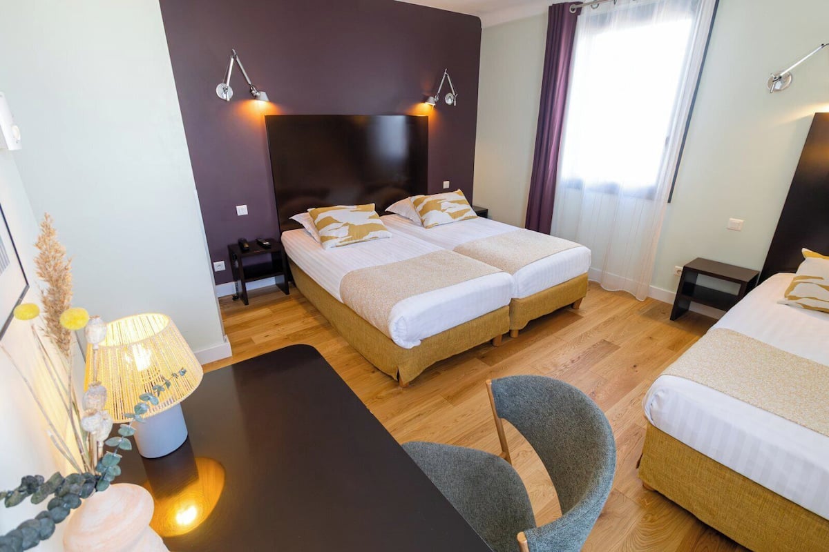 A hotel room with two beds, a desk, and a chair. There are bedside lamps and a small table with decorative items.