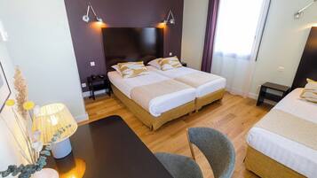 A hotel room with two beds, a desk, and a chair. There are bedside lamps and a small table with decorative items.
