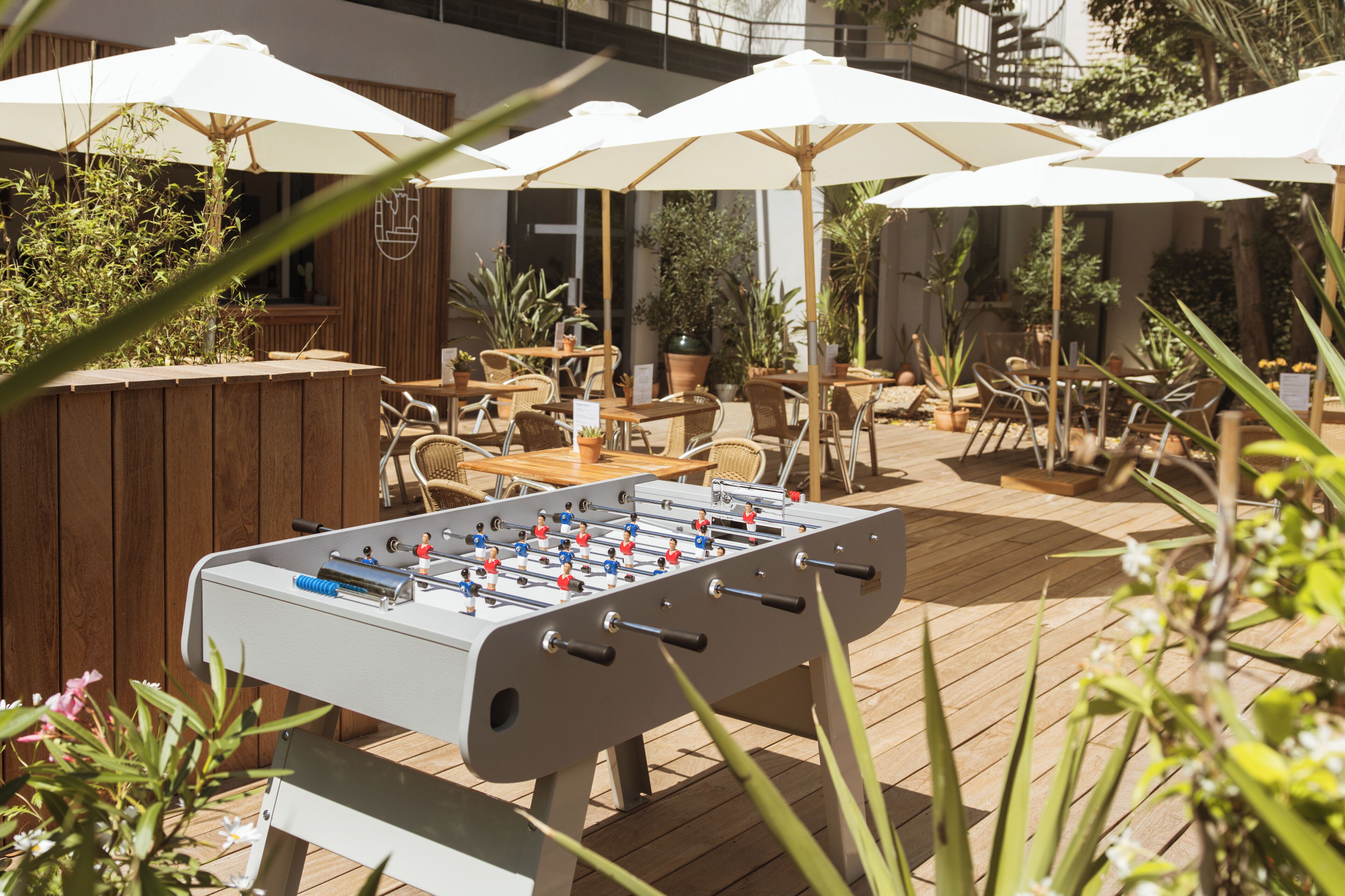 Un patio al aire libre con mesa de fútbol de mesa, mesas y sillas de madera, y sombrillas.