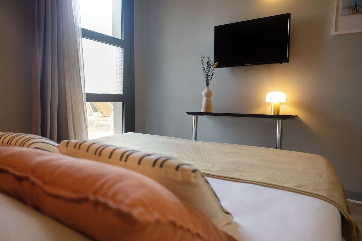 A hotel room with a bed, a TV mounted on the wall, a bedside table with a lamp, and a window with curtains.