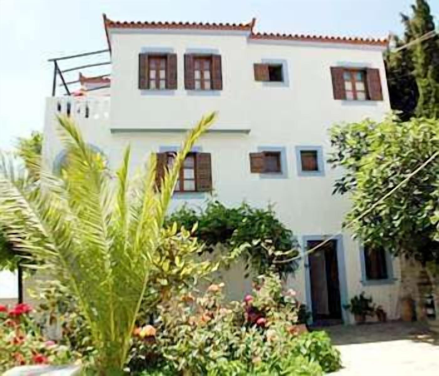 Un edificio de dos plantas con fachada blanca y contraventanas marrones, rodeado de follaje y flores.