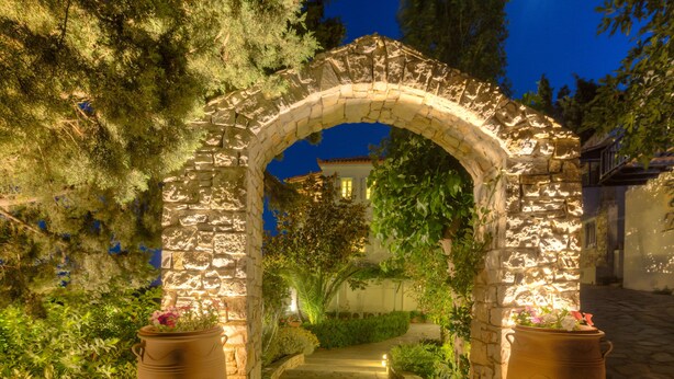 Un arco de piedra con escaleras que conducen a un edificio, rodeado de vegetación exuberante y plantas en macetas.