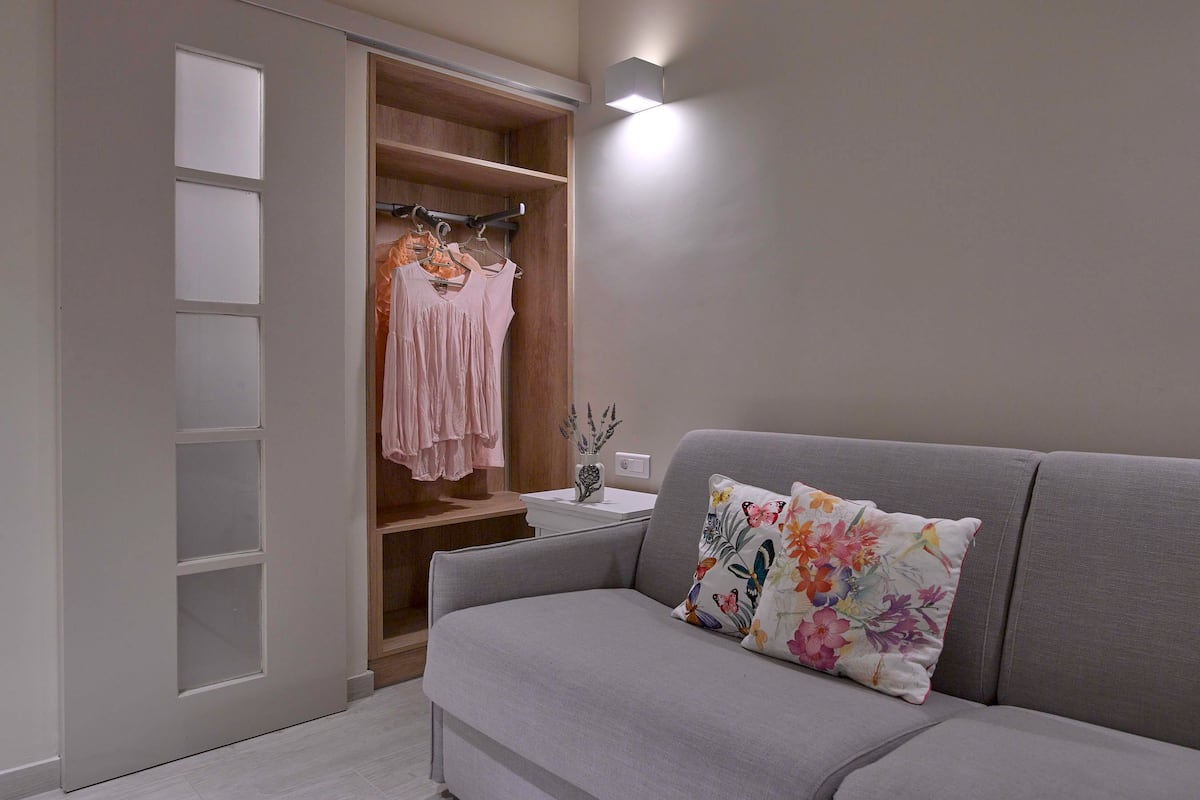 A modern living room with a grey sofa, a floral pillow, and a wooden wardrobe with a hanging rack.