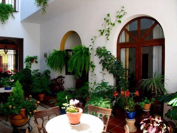 Um pátio com uma mesa redonda e cadeiras, cercado por plantas em vasos e uma porta de madeira.