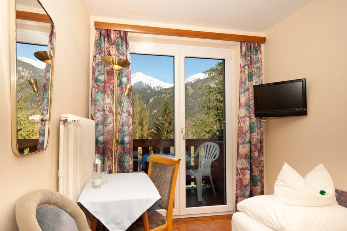 Ein Zimmer mit einem Bett, einem Tisch, einem Stuhl, einem an der Wand befestigten Fernseher und Bergblick durch eine Schiebetür.