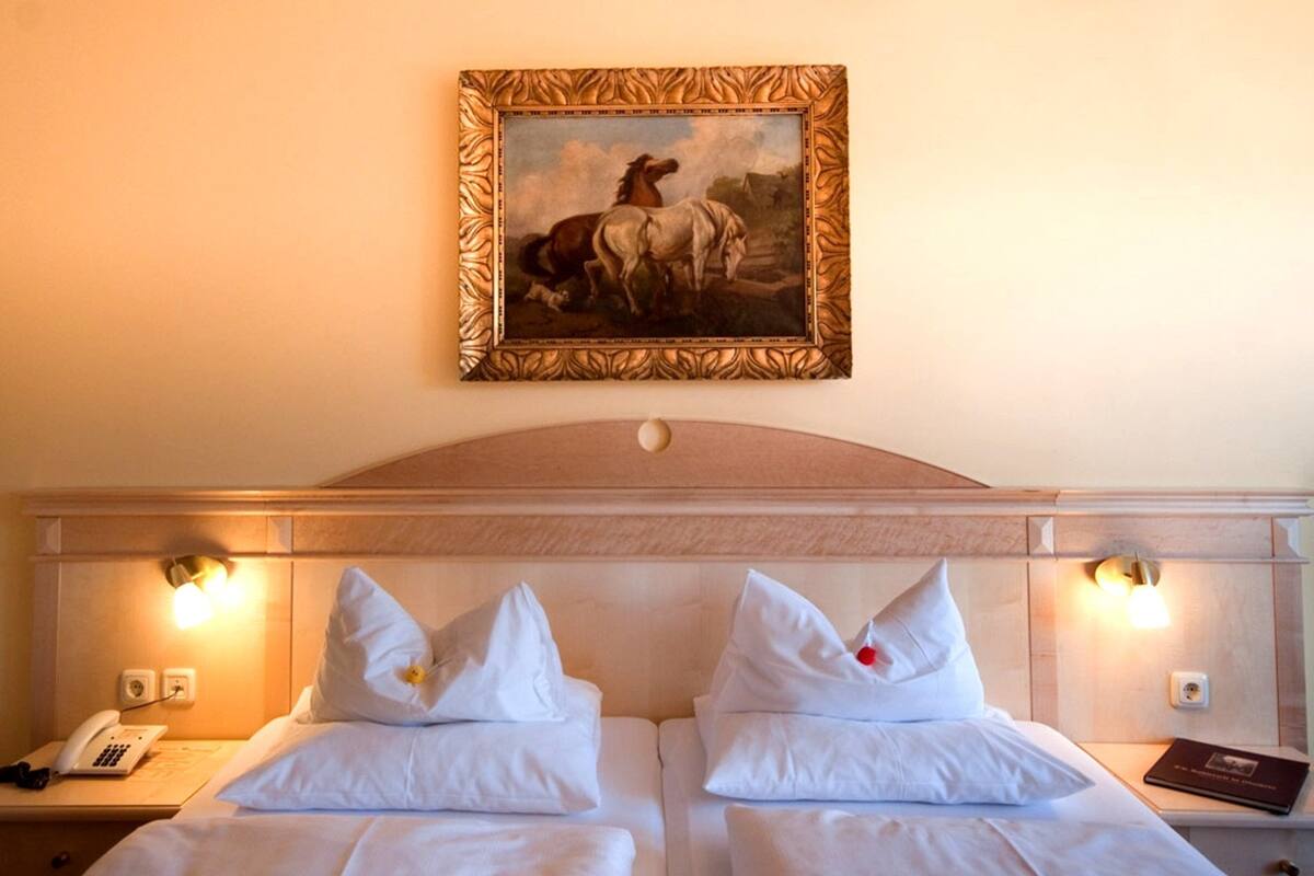 Ein Hotelzimmer mit zwei Betten, einem hölzernen Kopfteil, Nachttischlampen, einem Gemälde über den Betten, einem Telefon auf dem Nachttisch und einem Buch auf dem Nachttisch.