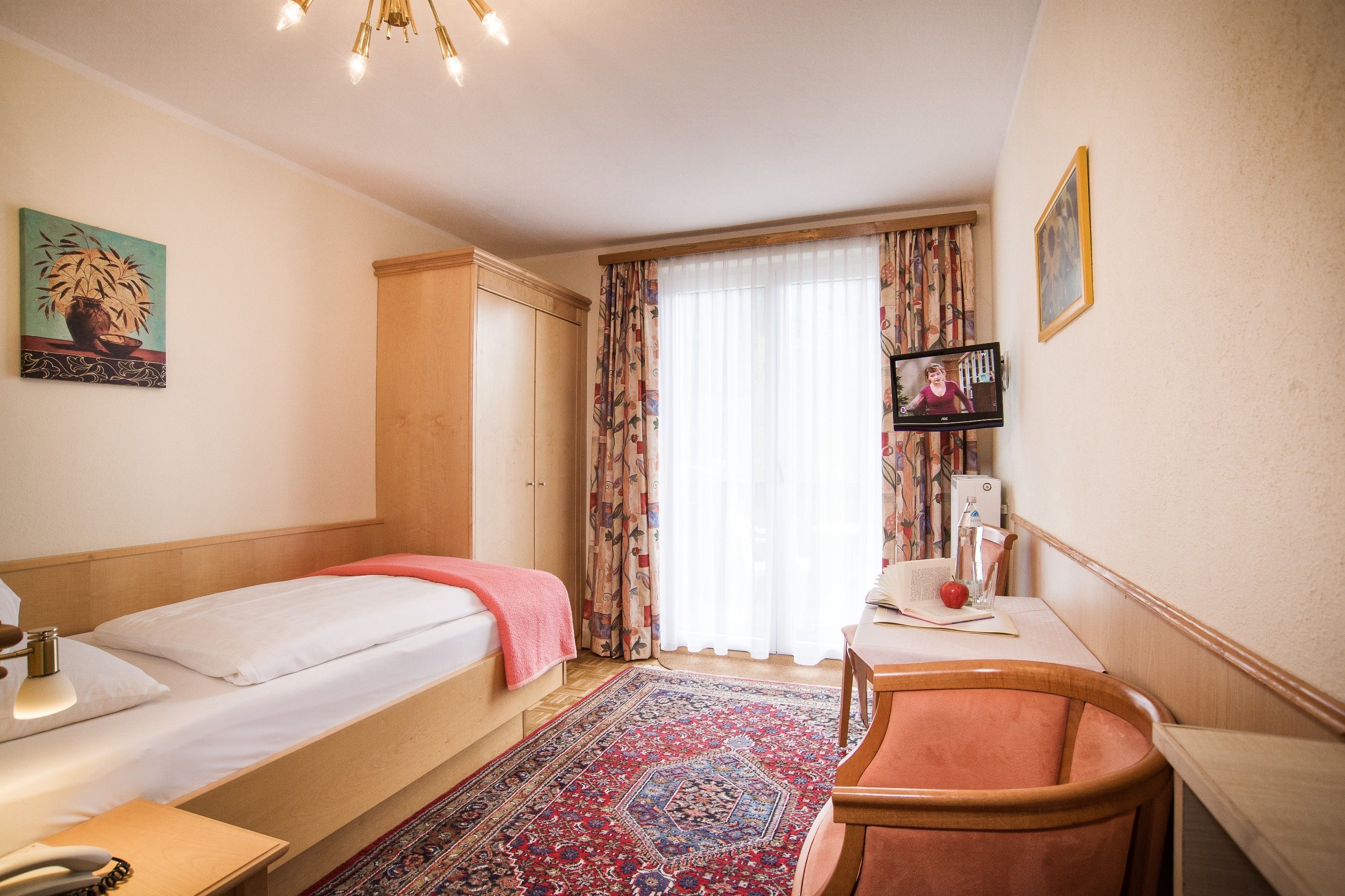 A hotel room with two single beds, a desk with a chair, a TV, and a large window with curtains.