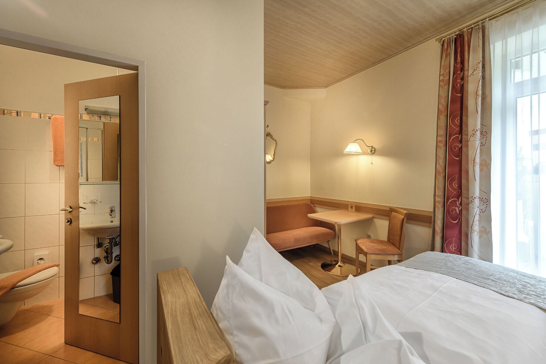 A hotel room with a bed, a bathroom with a sink and mirror, a small table, and a window with curtains.