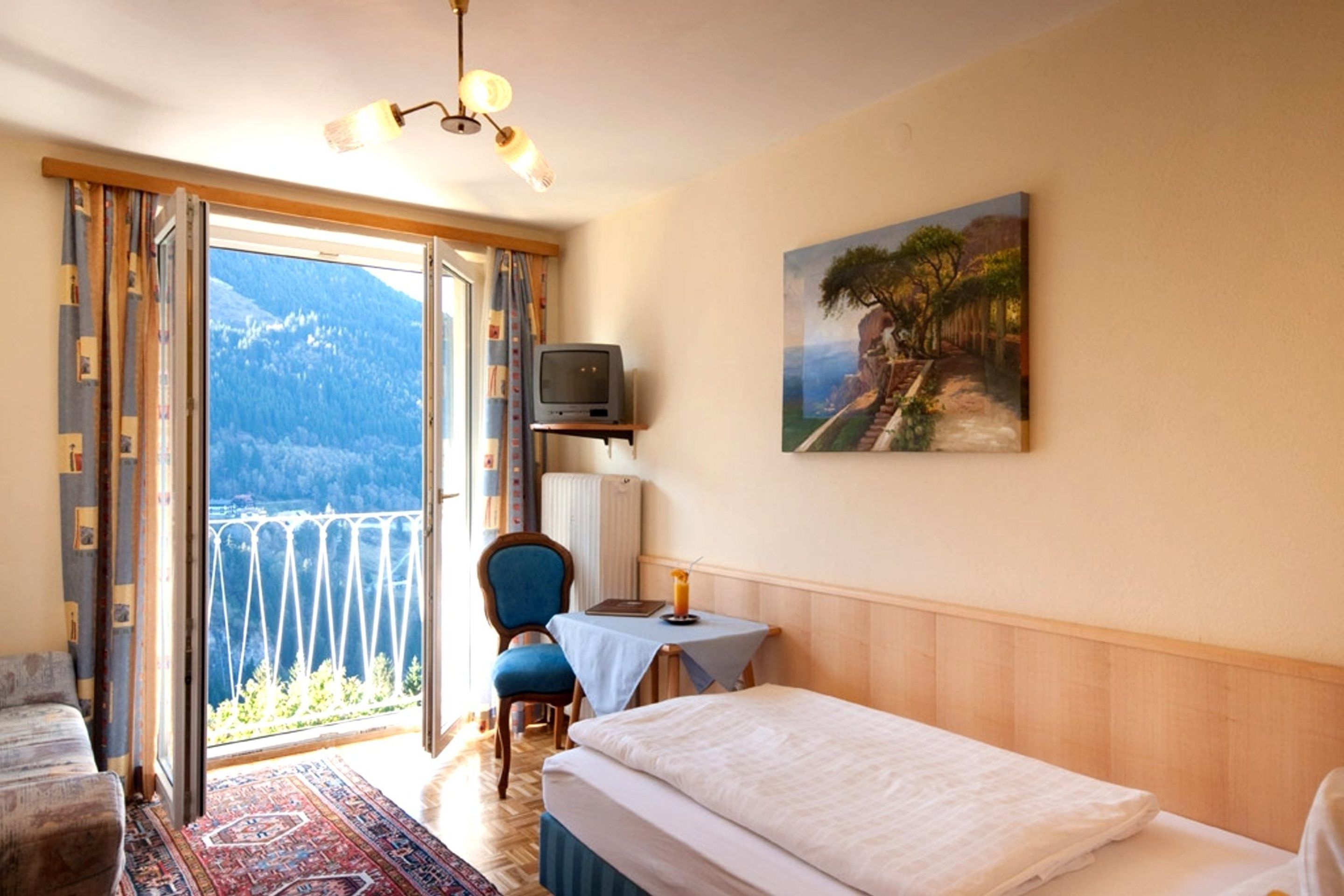 Ein Hotelzimmer mit Bett, Schreibtisch, Stuhl, einem Gemälde und Blick auf Berge durch die geöffnete Tür.