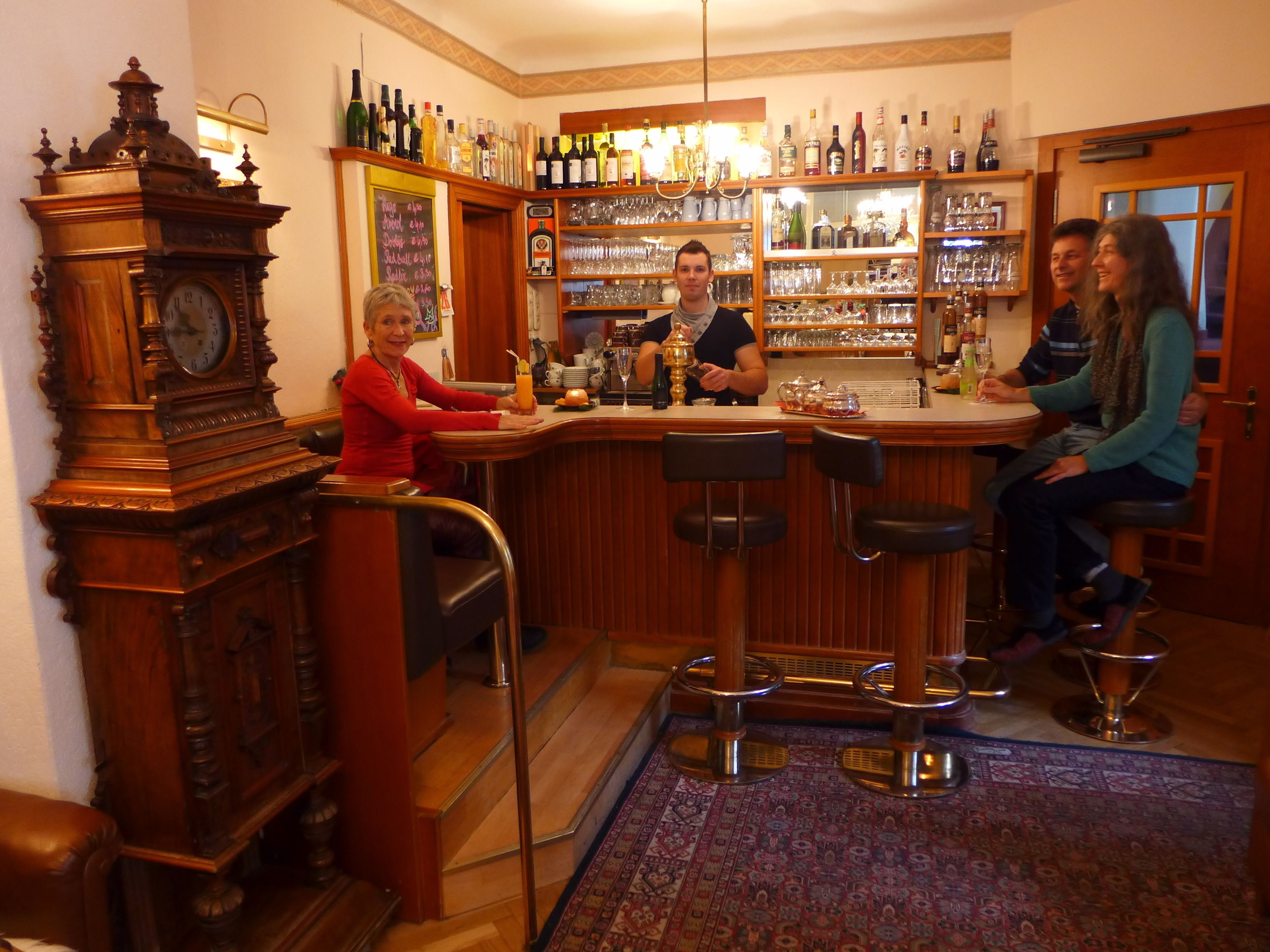 A bar area with a wooden bar counter, stools, a clock, and shelves with bottles and glasses.