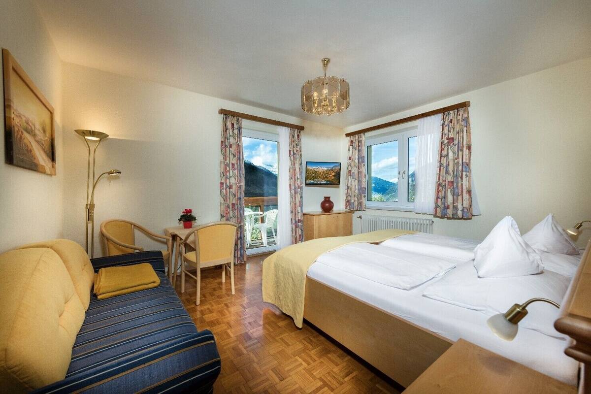 Ein Hotelzimmer mit Bett, einer Couch, einem Sessel, einem kleinen Tisch, einer Zimmerpflanze und Blick auf Berge durch das Fenster.