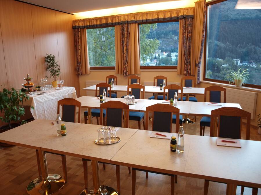 Un comedor con varias mesas y sillas, ventanas amplias con cortinas, y vista a un área boscosa.