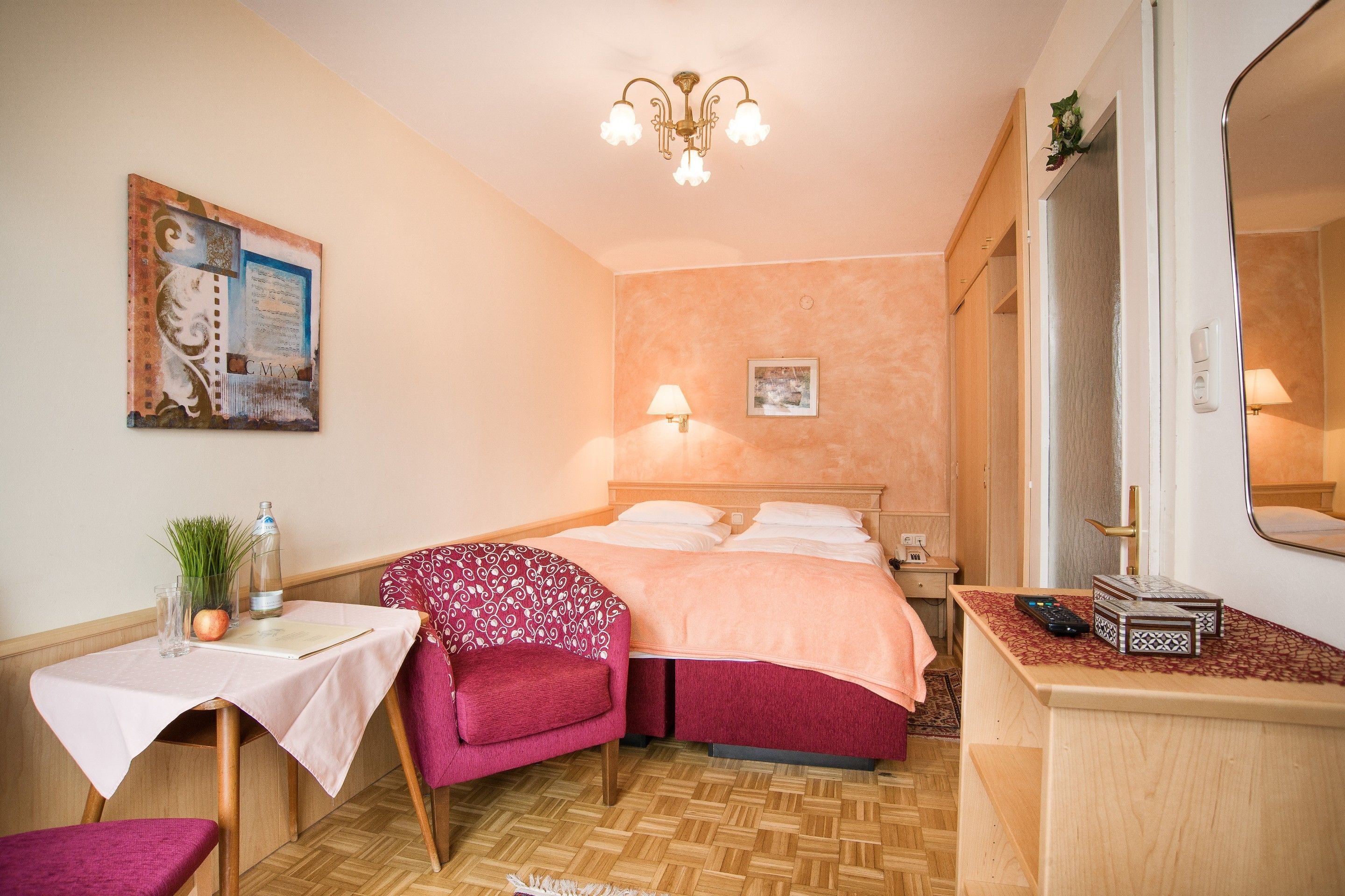 Hotellihuone, jossa on sänky, työpöytä tuolilla, pieni pöytä, peili ja kattokruunu.