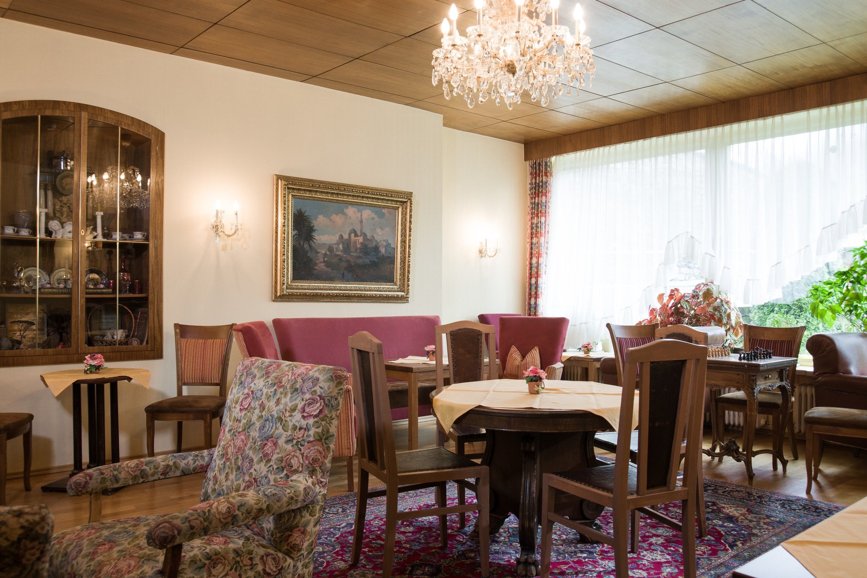 A dining area with a round table, chairs, and a chandelier. There is a floral armchair and a cushioned bench. A painting is on the wall, and a window with curtains is visible.