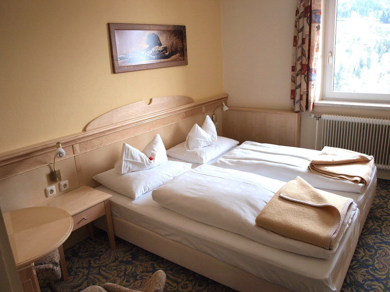 Habitación de hotel con dos camas, un escritorio pequeño, una mesita de noche, un cuadro en la pared y una ventana con cortinas.