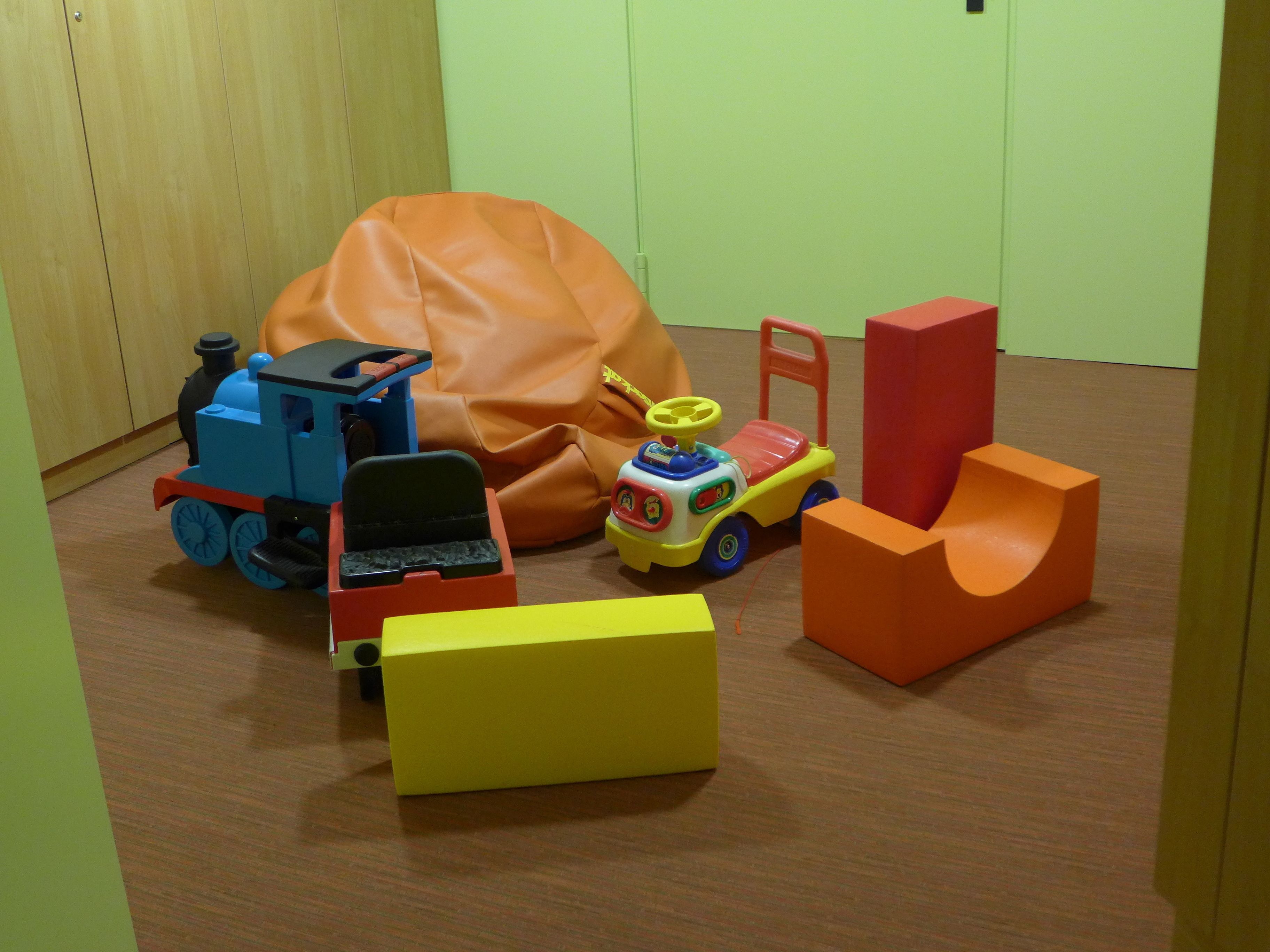 A room with a wooden floor, a green wall, and a wooden wardrobe. There are colorful toy cars and a yellow box on the floor.