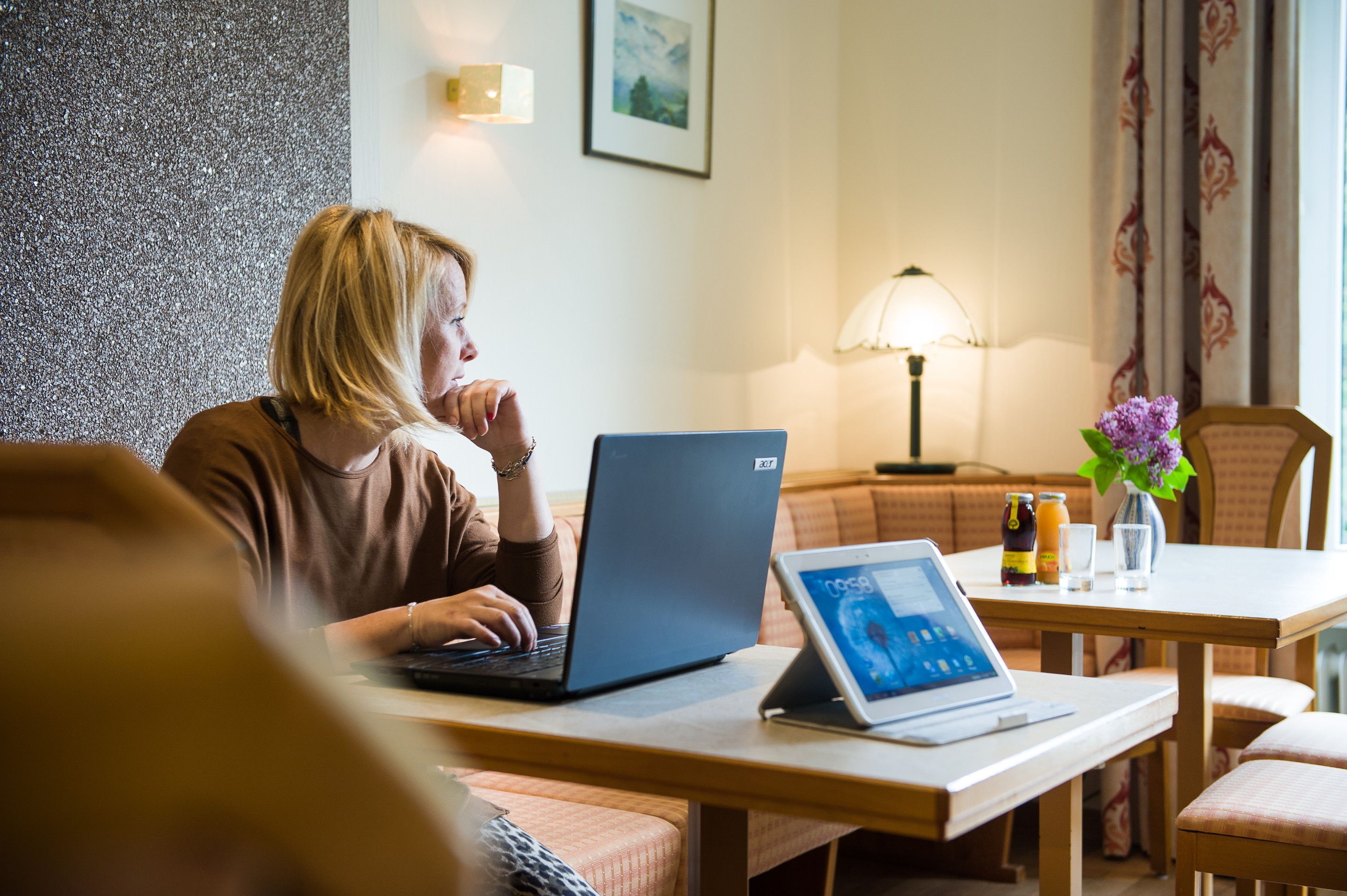 A person using a laptop at a table in a room with a sofa, a lamp, and a vase with flowers.