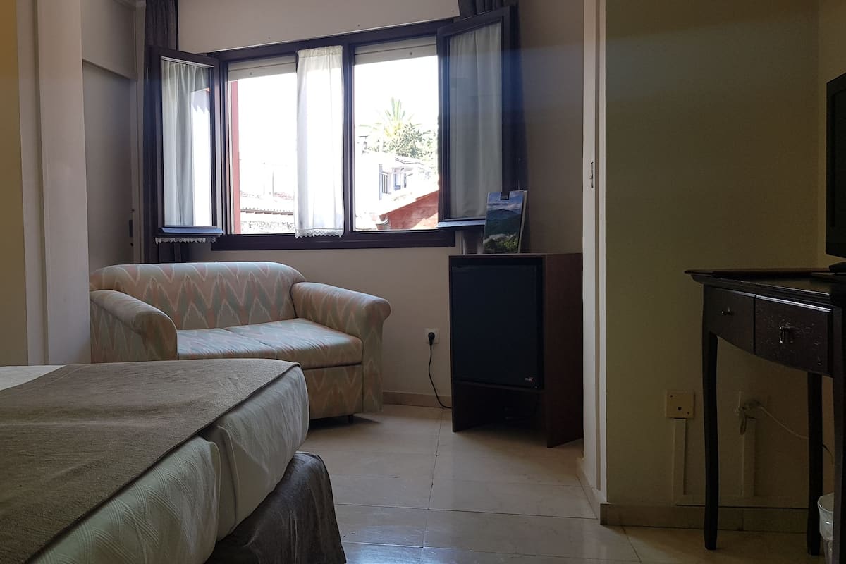 Quarto de hotel com duas camas, uma poltrona pequena, uma televisão e uma janela com cortinas.