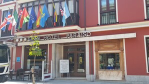 A hotel facade with multiple flags, a signboard, and a vehicle parked outside. - Gran Hotel Paraiso (Llanes)