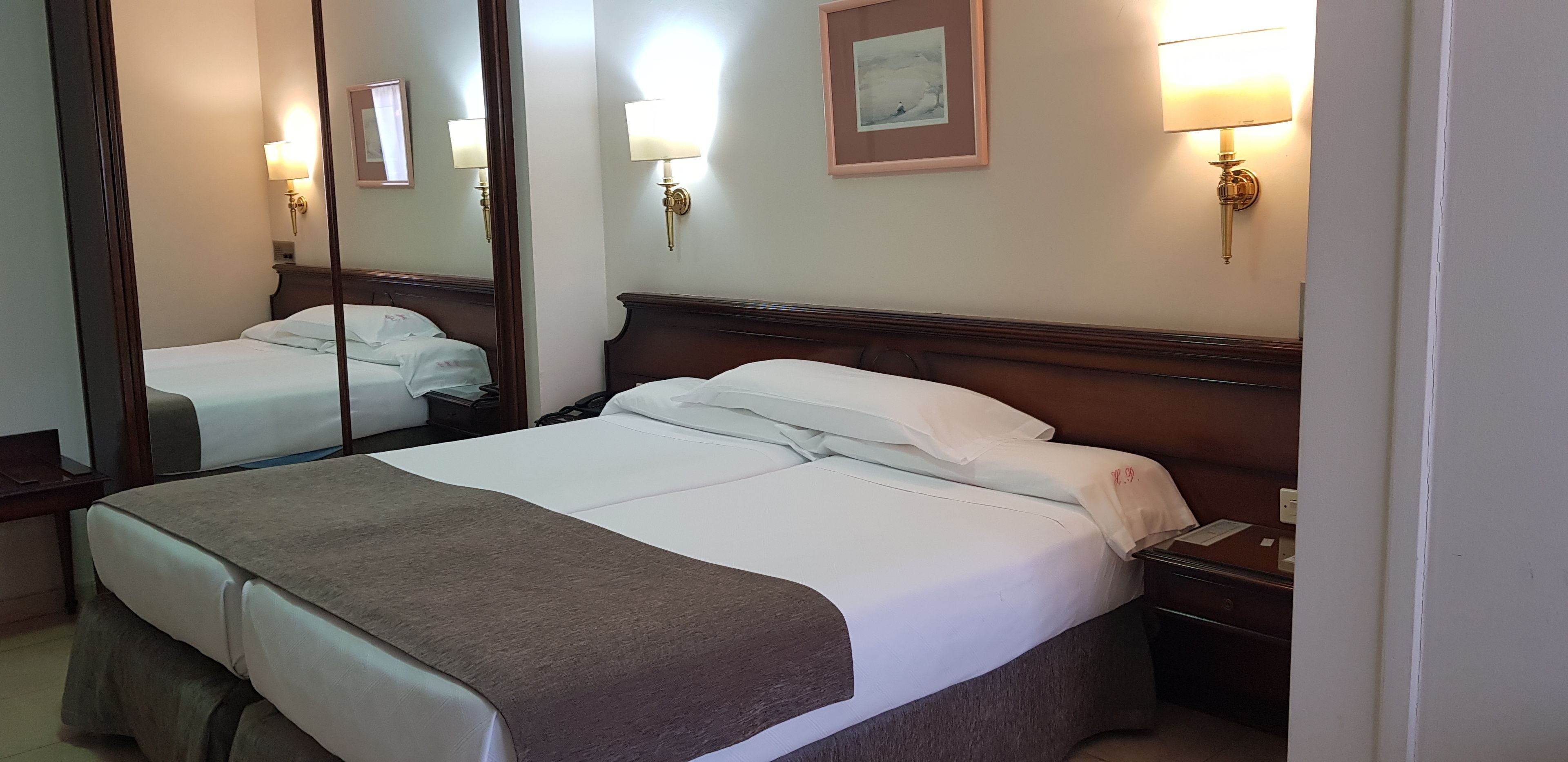 Ett hotellrum med en stor säng, ett sängbord, en nattdukslampa, en spegel och väggmonterade lampor.