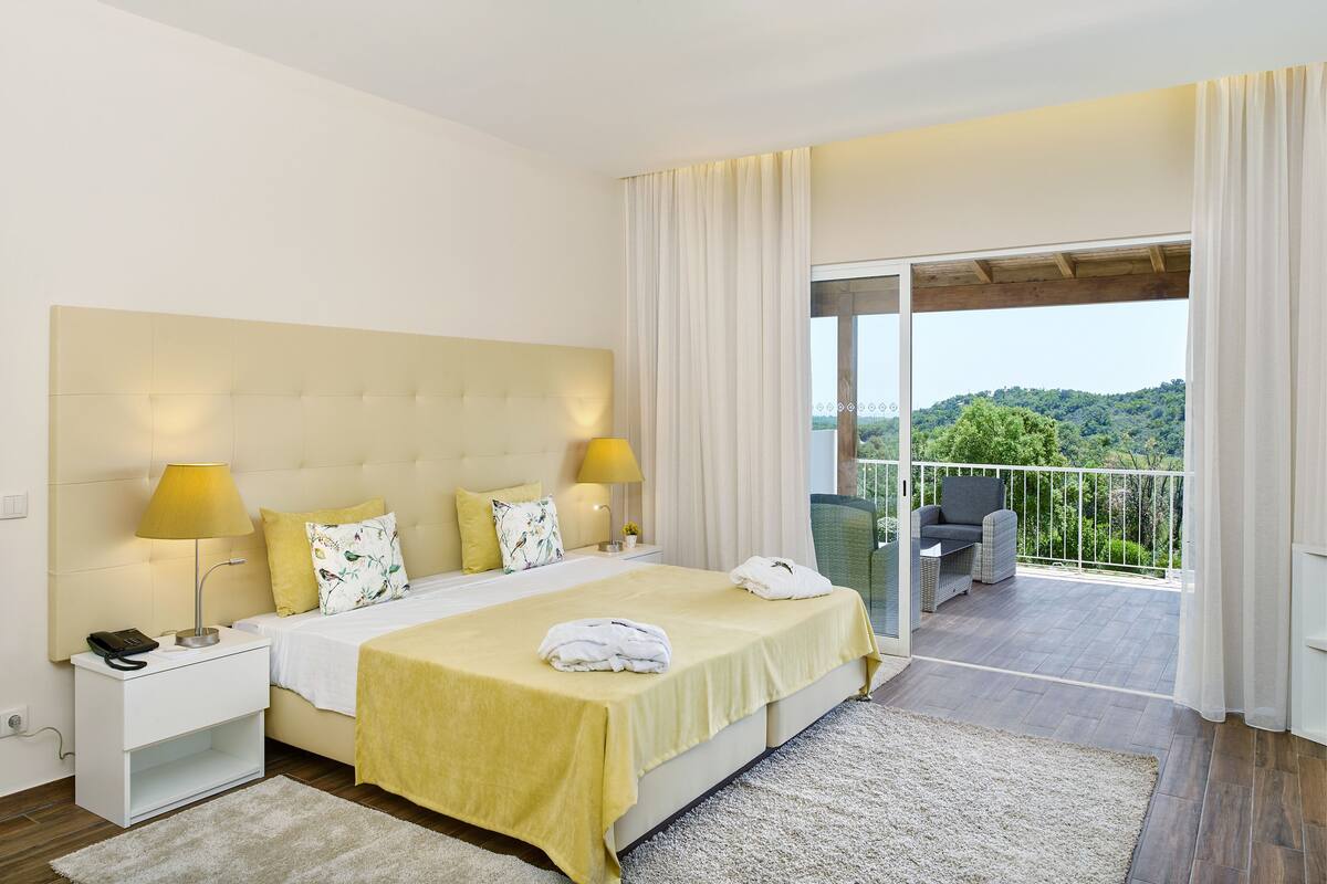 A hotel room with a large bed, two bedside lamps, a nightstand, a balcony with a view of the hills.