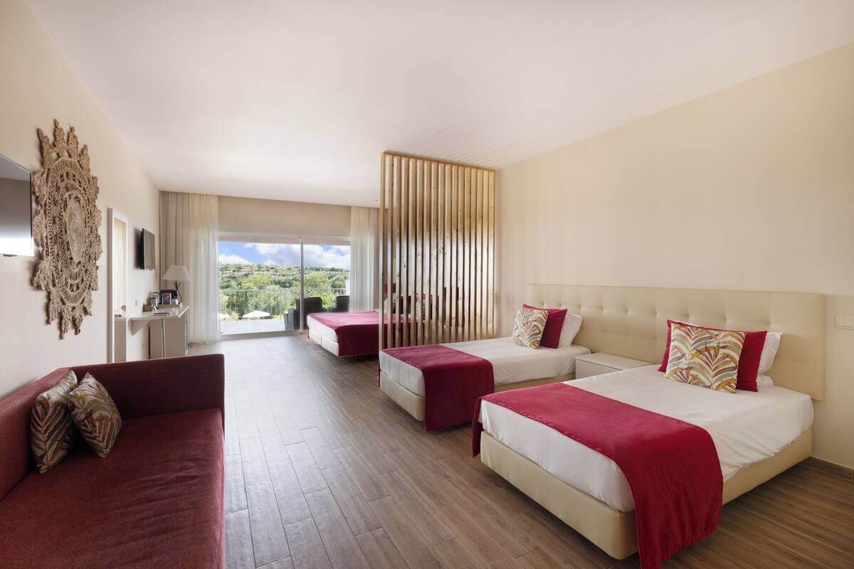 A hotel room with two beds, a sofa, a TV, and a large window with a view of greenery.