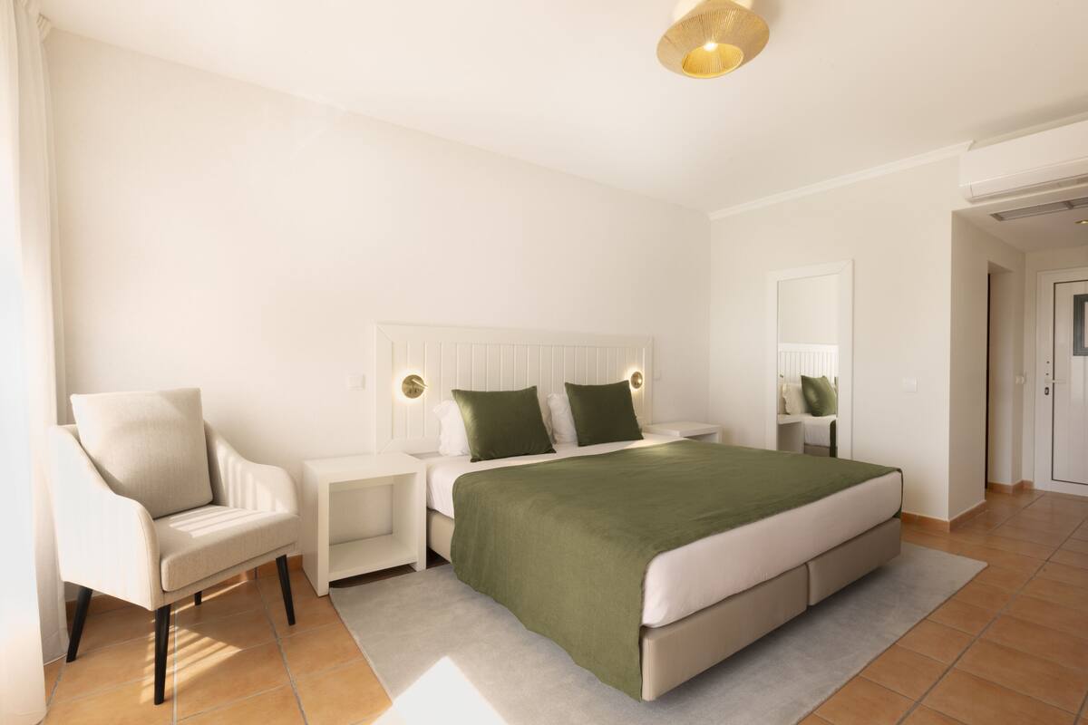 A modern bedroom with a large bed, a chair, a nightstand, and a bedside table.