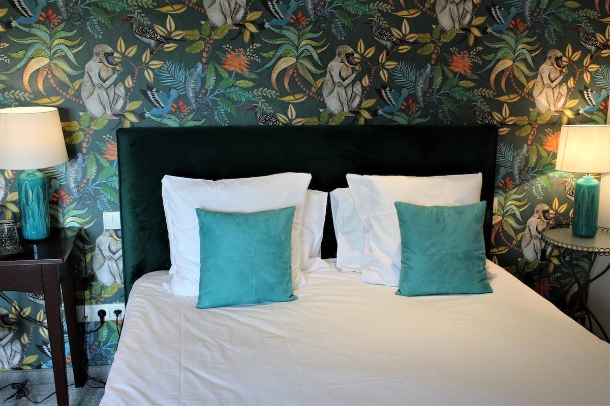 A neatly made bed with white linens and two teal pillows, flanked by two bedside tables with lamps, against a wall with a detailed floral and animal mural.