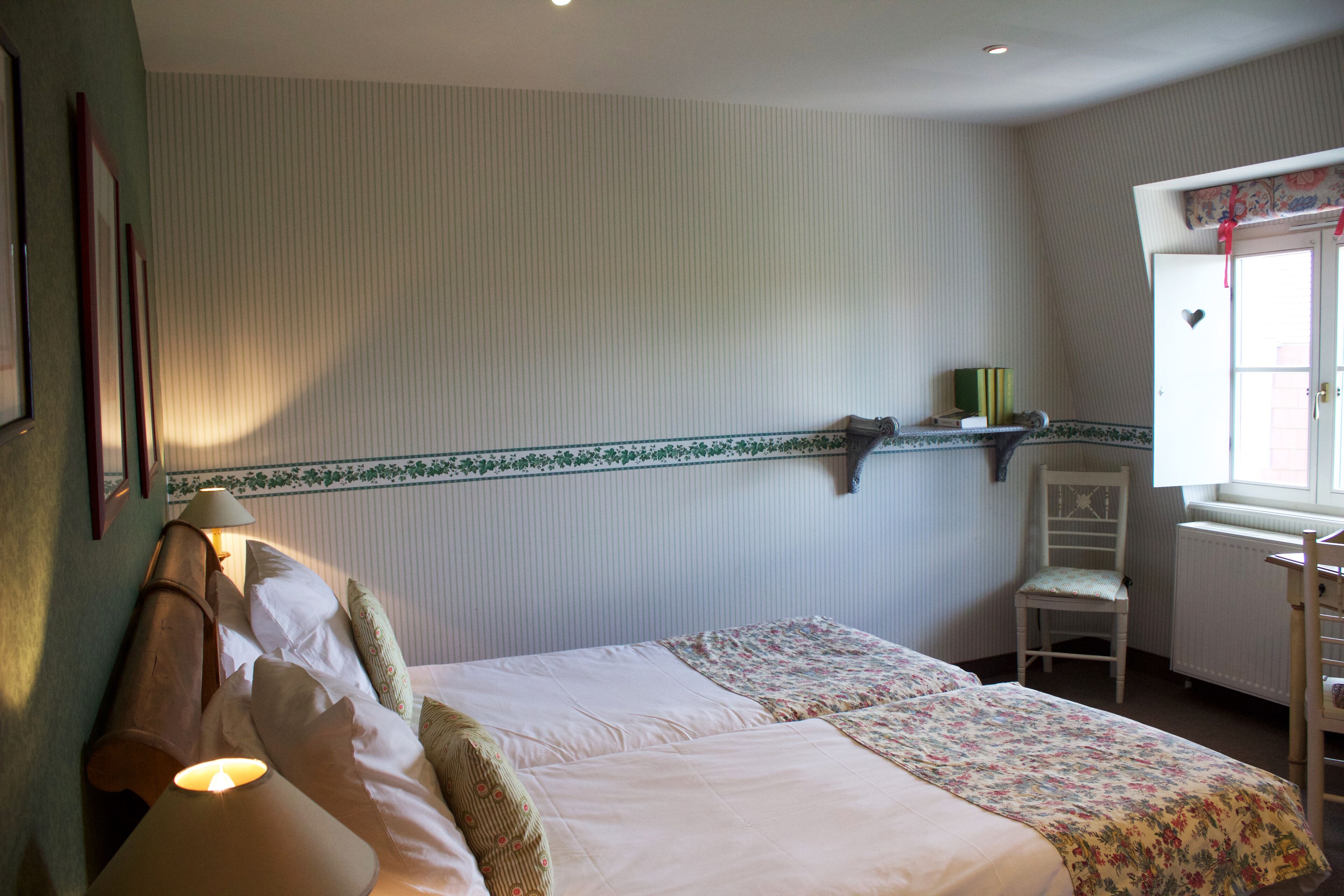A bedroom with two beds, a chair, a shelf with books, and a window with a view.