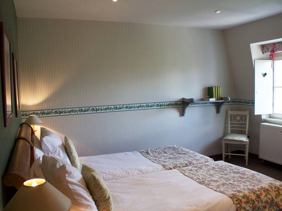 A bedroom with two beds, a chair, a shelf with books, and a window with a view.