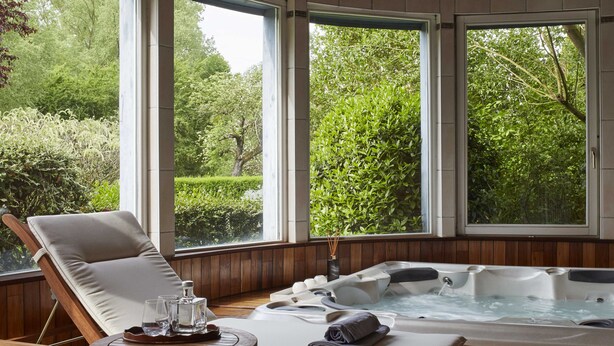 Un área de spa con un sillón de madera, un jacuzzi y vistas a la vegetación exuberante a través de amplios ventanales.
