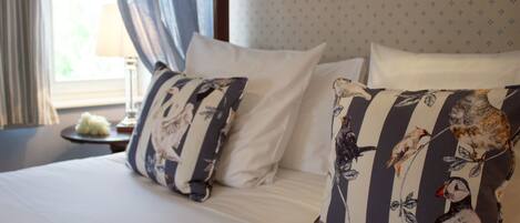 Una cama bien hecha con almohadas estampadas con motivos florales y una almohada lisa, una mesita de noche con una lámpara y una ventana con cortinas.