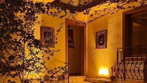 Un espacio exterior con cálida iluminación, escalones de piedra que conducen a una puerta, y macetas con plantas junto a un pequeño jardín.