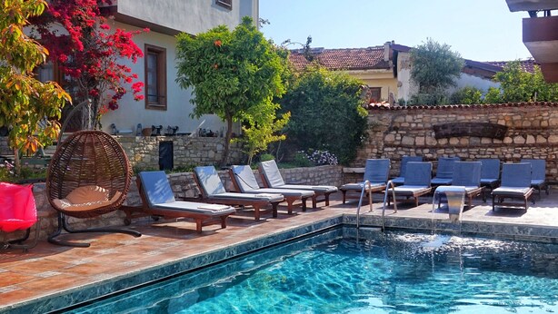 Una piscina cristalina con sillones de descanso y una silla de mimbre, rodeada por un muro de piedra y un edificio con techo de tejas.