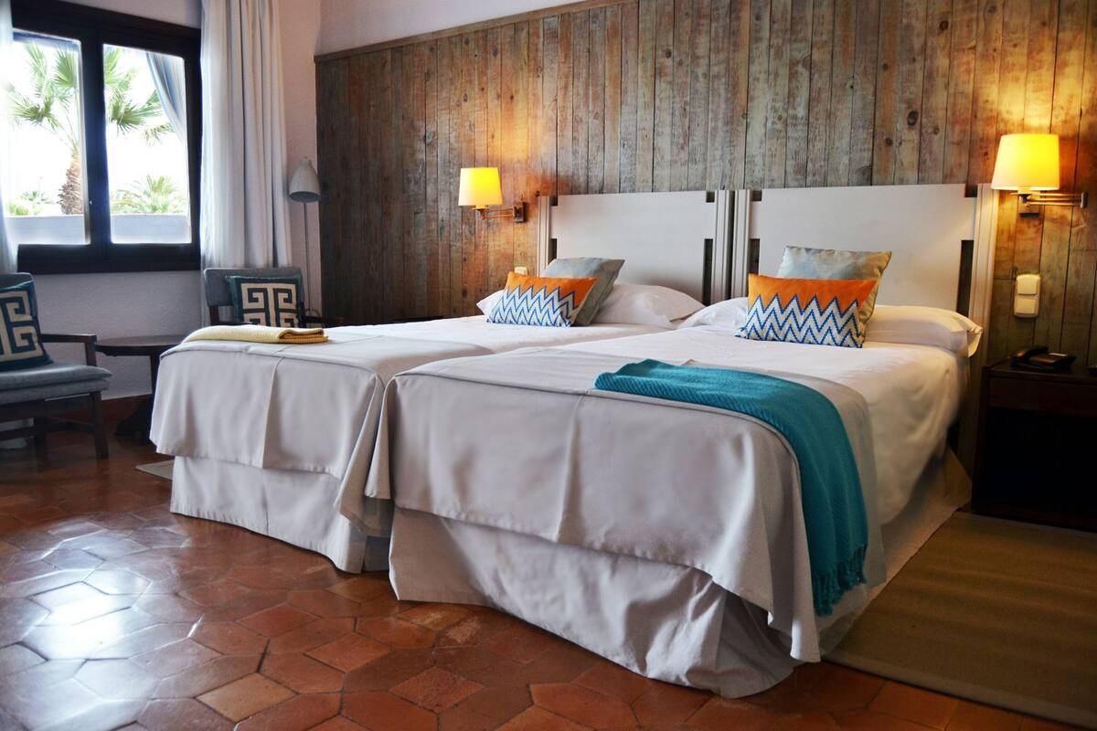 Une chambre d’hôtel avec deux lits, un mur en bois, une fenêtre avec des rideaux, une chaise et une lampe.