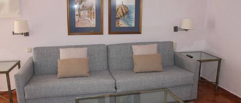 Una sala de estar con un sofá gris, dos cuadros enmarcados en la pared, una mesa de centro de vidrio y dos mesitas de noche con lámparas.