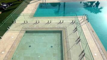 Una gran piscina al aire libre con una barrera de cristal, una piscina más pequeña y sillas de descanso.
