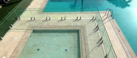 Une grande piscine extérieure avec une paroi en verre, une piscine plus petite et des chaises longues.