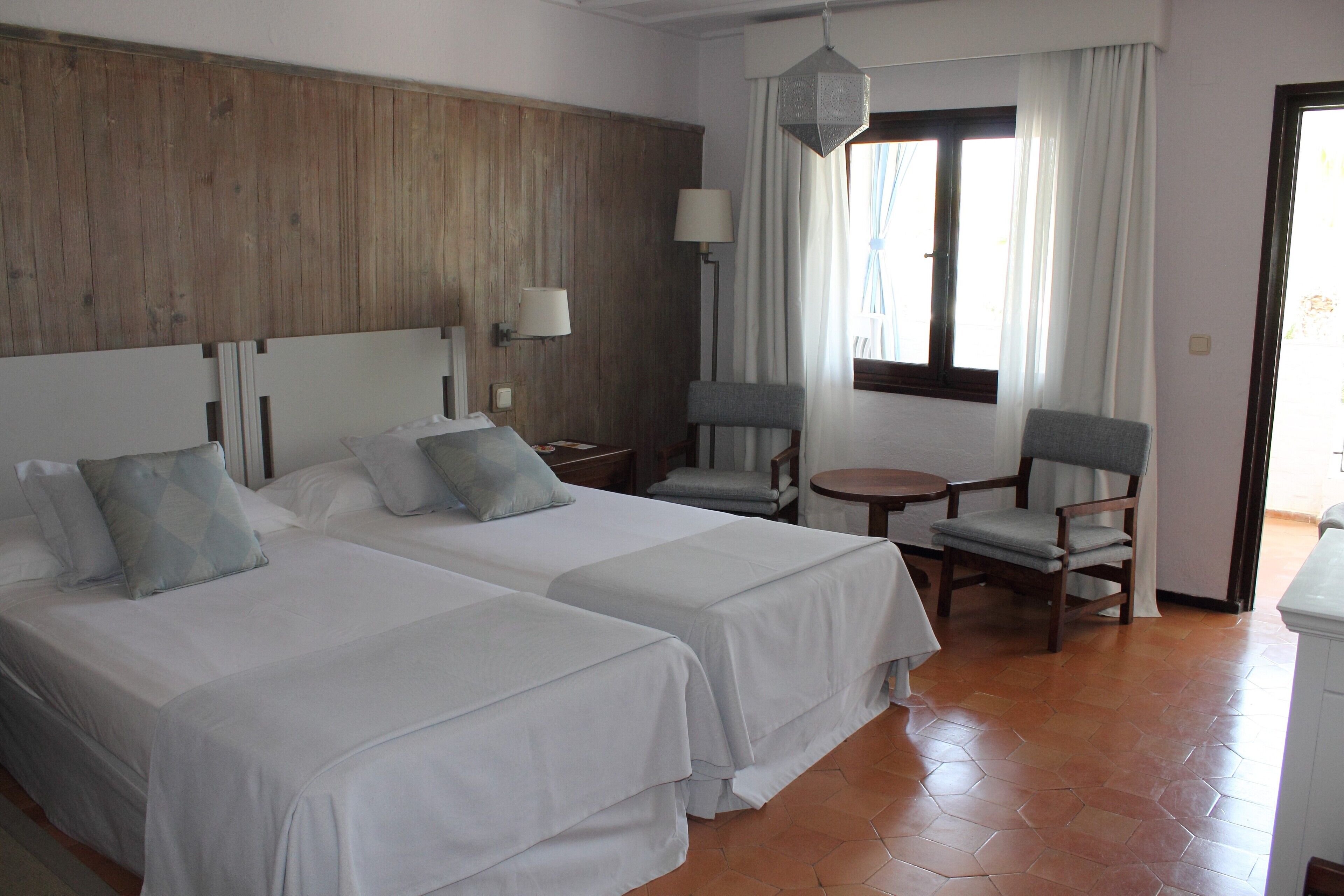 Habitación de hotel con dos camas, una mesita, dos sillas, una lámpara y un ventanal con cortinas.
