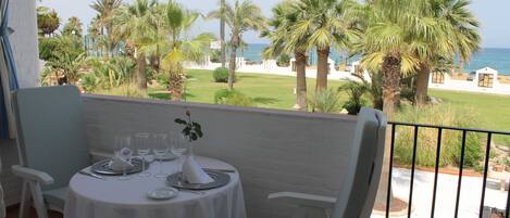 A balcony with a table set for two, overlooking a beach with palm trees and a clear sky.