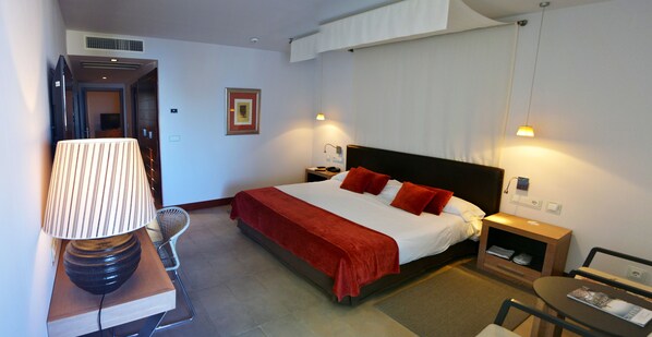 Habitación de hotel con cama, mesitas de noche, lámpara, una silla y una pequeña mesa con una revista.