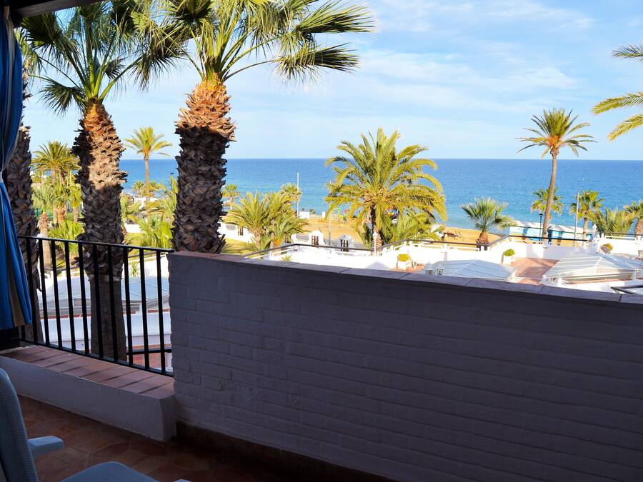 A balcony with a view of the sea, palm trees, and beach umbrellas.