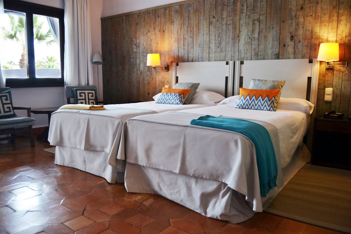 Une chambre d’hôtel avec deux lits, des lambris en bois aux murs, une fenêtre donnant sur des palmiers et un sol carrelé.