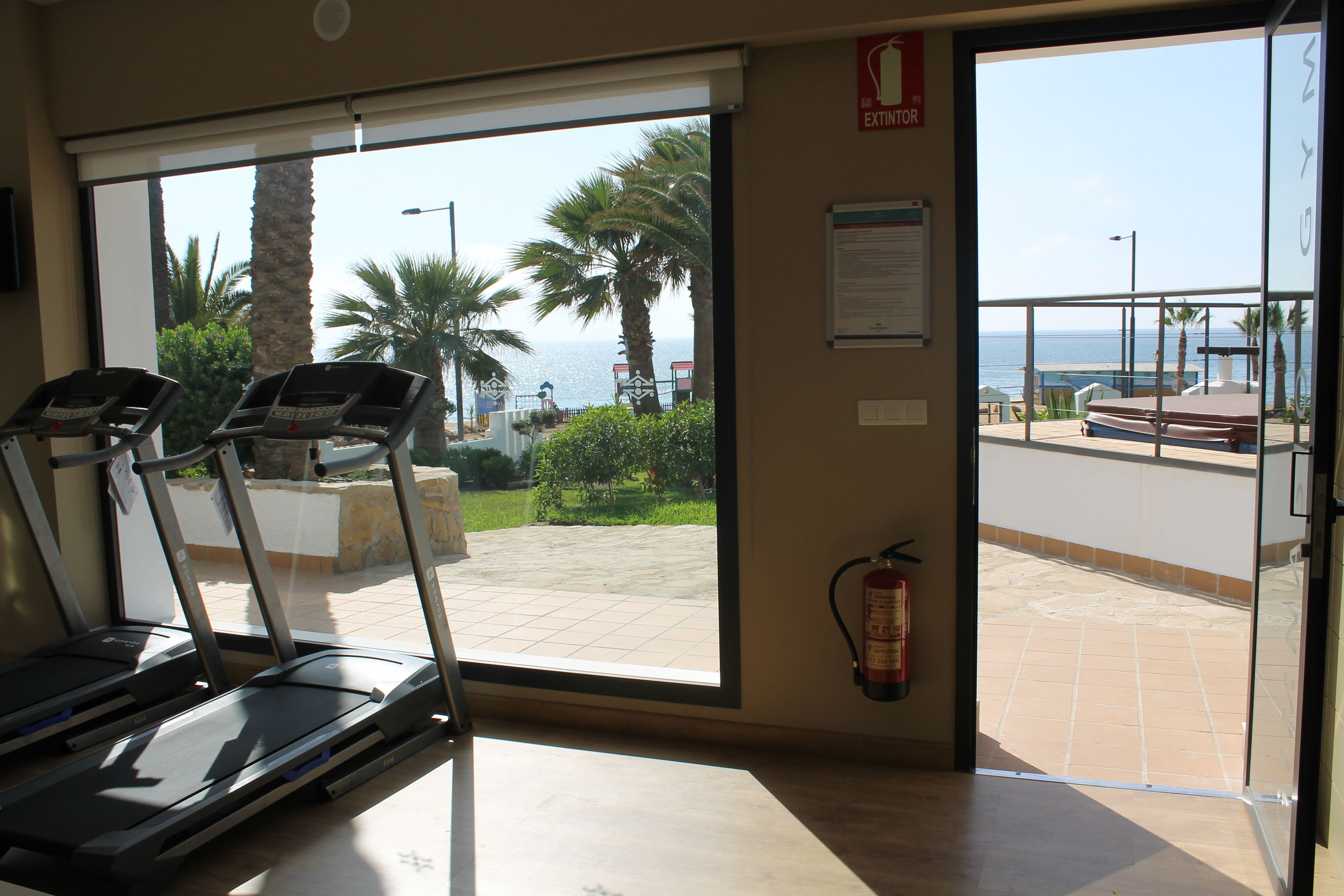 A room with a treadmill, a fire extinguisher, and a view of the sea through a glass door.